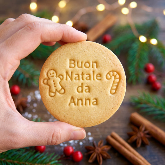 Biscotto rotondo tenuto in mano con scritta Buon Natale da Anna, omino di pan di zenzero e bastoncino di zucchero.
