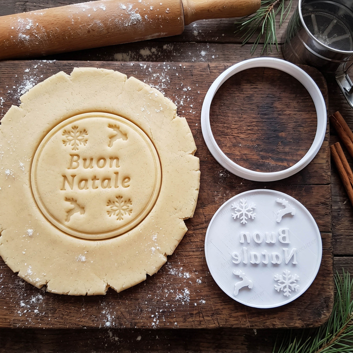 Tagliabiscotti rotondo e timbro Buon Natale con renne e fiocchi di neve, accanto alla frolla appena stampata.
