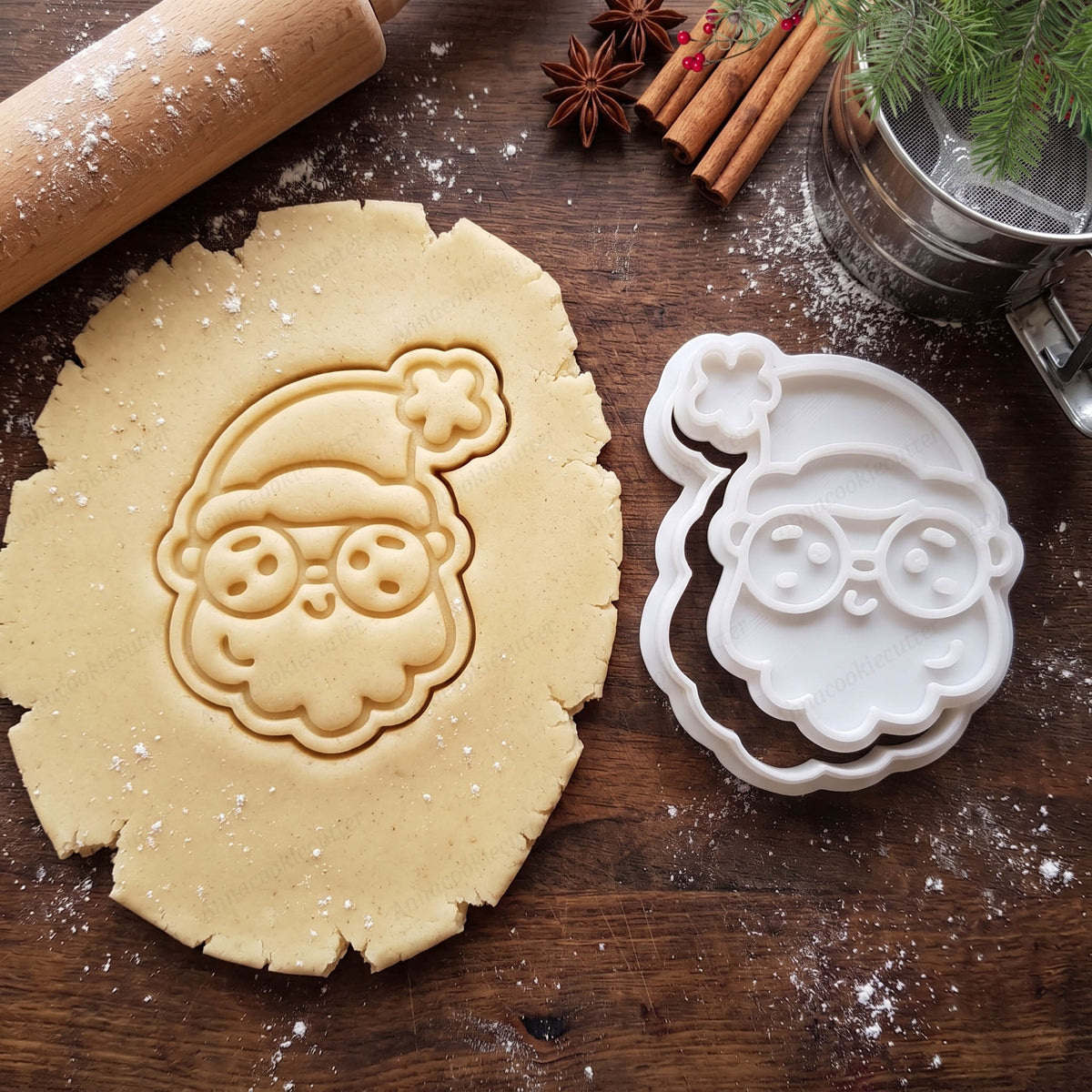 Viso di Babbo Natale con occhiali impresso nella pasta frolla, con tagliabiscotti e timbro bianchi accanto.
