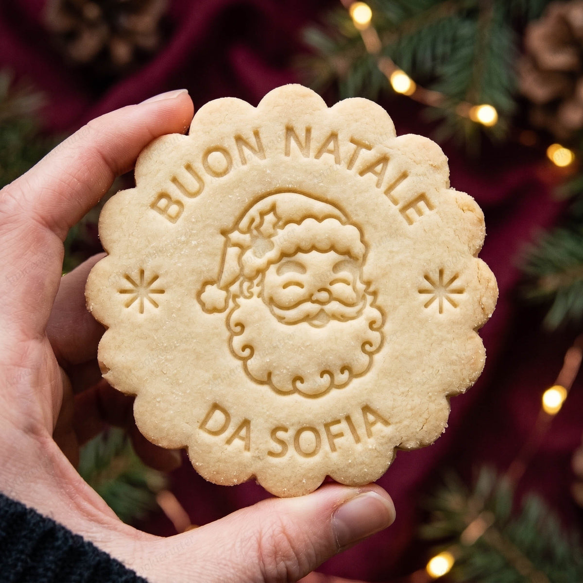 Biscotto dal bordo festonato con volto di Babbo Natale e scritte Buon Natale e Da Sofia, tenuto in mano.
