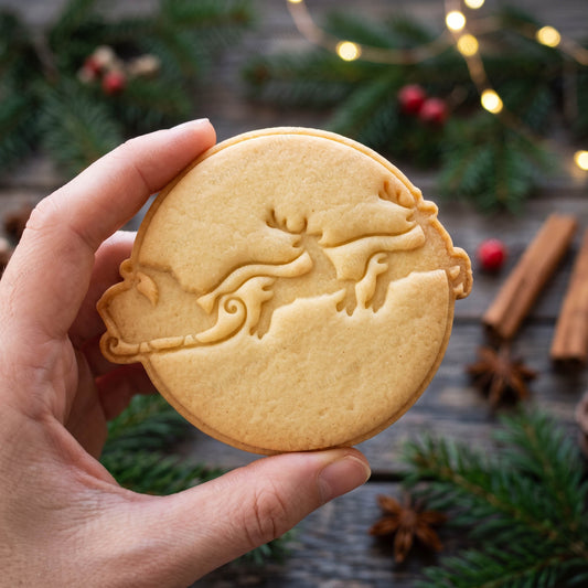 Biscotto rotondo tenuto in mano con la slitta di Babbo Natale trainata da due renne impressa sulla superficie.
