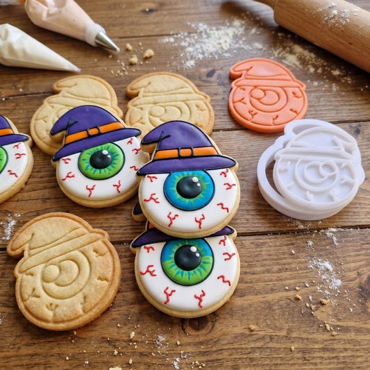 Biscotti Halloween decorati a forma di occhio con cappello da strega, accanto a timbro e tagliabiscotti.