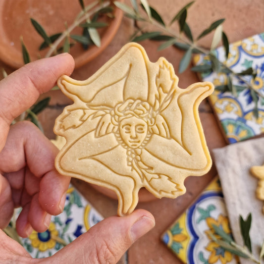 Biscotto a forma di Trinacria siciliana con volto centrale, tre gambe e spighe, tenuto in mano.
